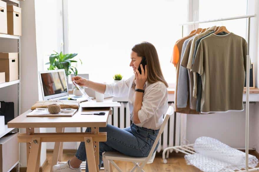 A woman working in a home office, talking on the phone while using a laptop, with clothing on a rack in the background.