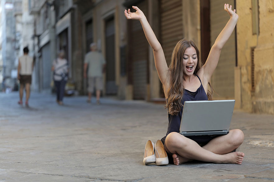 woman raising her hands sitting on a street with laptop