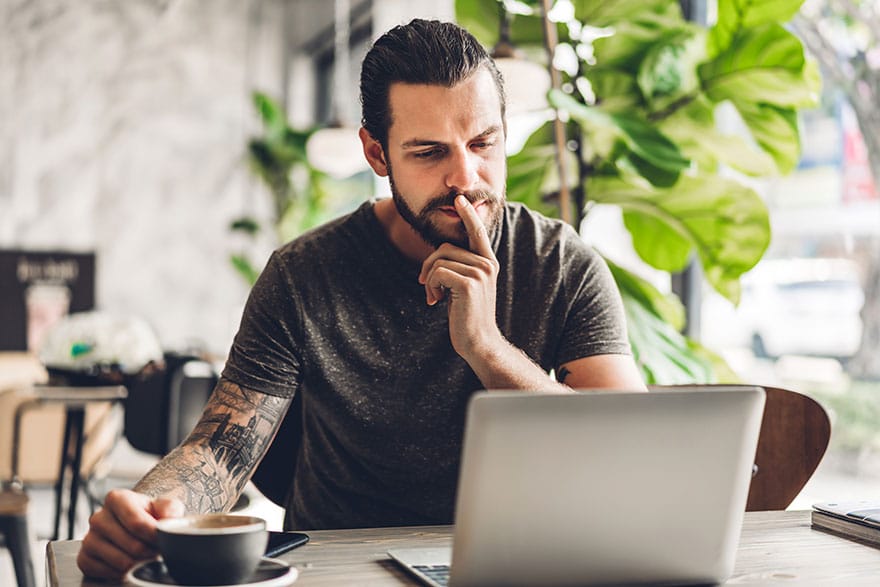 Picture of a man sitting behind a laptop with a finger on his chin to show that he is thinking