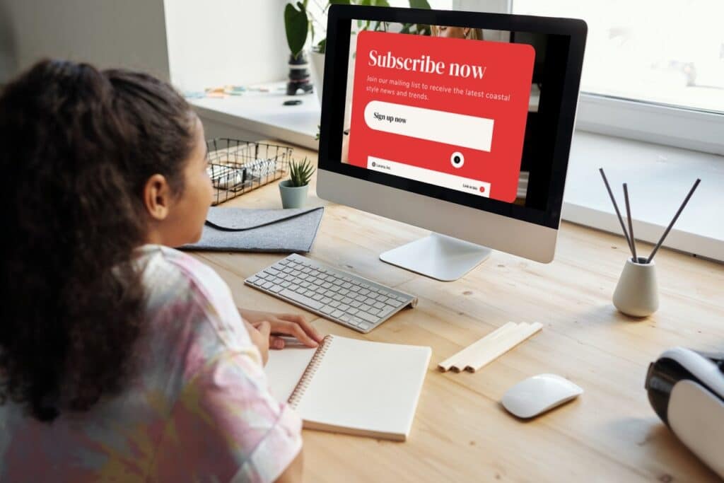 Woman sitting at a desktop with an empty notebook, on the desk is a keyboard and a monitor that shows a big red popup which reads "Subscribe now" to illustrate interstitial ads/popups which aren't good for web vitals. 