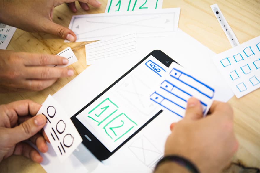 Two sets of hands holding paper cutouts of mobile elements in their hands like icons section, dropdowns, and text. On the table lies a full page with mobile as the background and logo with two column section to illustrate building user experience.