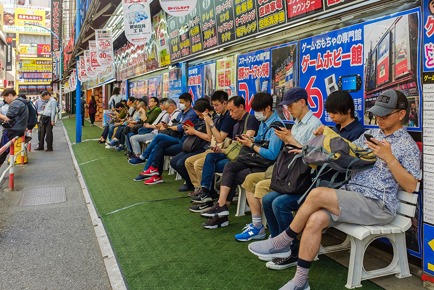 Several people seated on a very long bench looking at their mobile phones