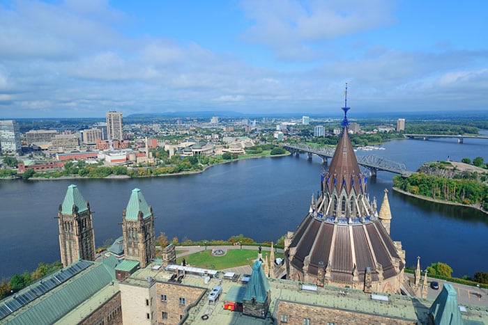 Aerial view of Ottawa, Canada, featuring the Library of Parliament with its distinctive pointed roof in the foreground. The Ottawa River winds through the scene, with bridges connecting to the Gatineau area in the background. The skyline includes a mix of modern buildings and natural greenery under a partly cloudy sky.