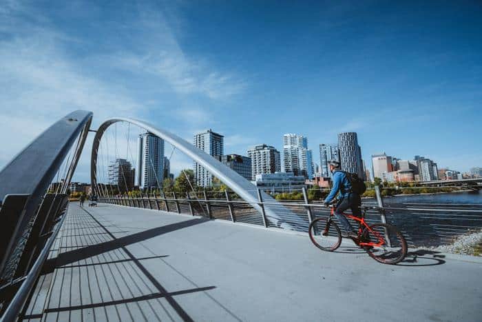 Cyclist riding on a modern pedestrian bridge in Calgary, with a view of the downtown skyline and the river below. The clear blue sky and contemporary architecture highlight Calgary's urban charm and outdoor lifestyle.