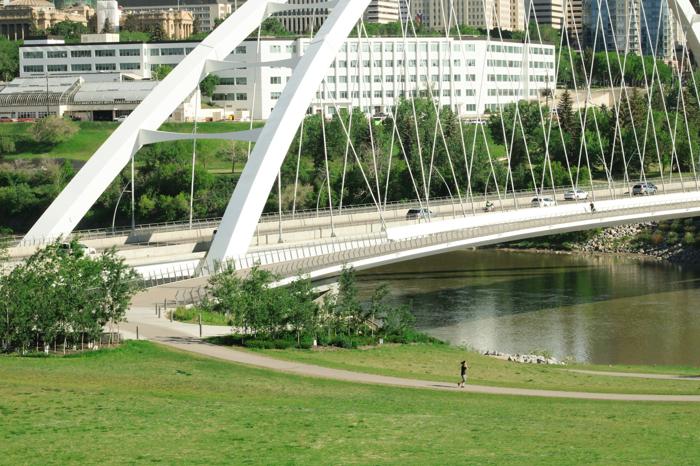 View of Edmonton's modern white bridge spanning over a calm river, surrounded by green spaces and city buildings in the background. A jogger is seen on a nearby pathway, showcasing the city's mix of urban infrastructure and natural landscapes.
