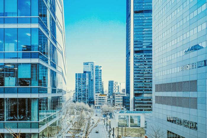 Modern downtown Montreal cityscape with high-rise buildings and glass facades reflecting a bright blue sky. A street lined with trees and urban structures highlights the vibrant and contemporary feel of the city.