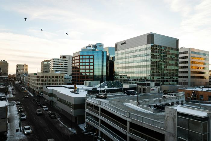 Downtown Regina cityscape with modern office buildings reflecting the evening light. Snow-covered rooftops and parked cars line the streets, while birds fly across the clear sky, capturing a peaceful winter scene.