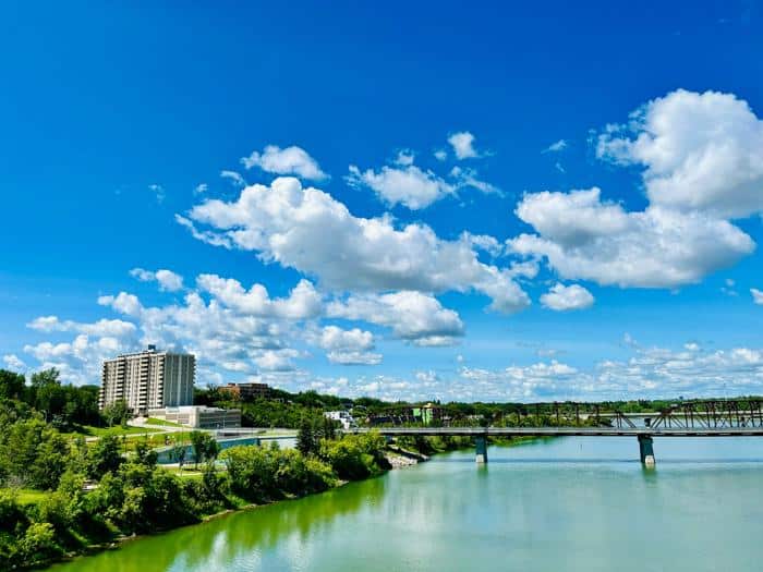 Scenic view of Saskatoon featuring a calm river, lush greenery, and a bridge under a bright blue sky with scattered clouds. Modern buildings and natural landscapes combine to highlight the city's serene and picturesque environment.