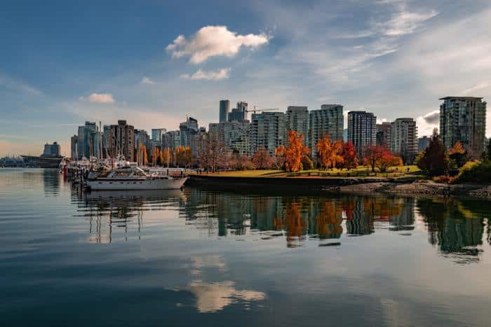 Scenic view of Vancouver's skyline reflecting on calm waterfront waters, surrounded by vibrant autumn trees and docked boats. The image captures the city's blend of urban and natural beauty under a partly cloudy sky.
