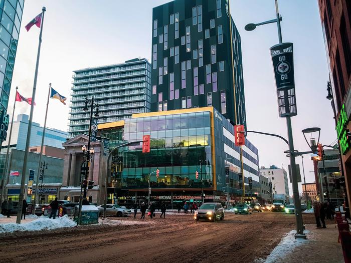Downtown Winnipeg street scene with modern glass buildings, snowy roads, and pedestrians crossing at the intersection. The cityscape features a mix of urban activity and architecture, with Canadian flags and street banners adding to the vibrant winter setting.