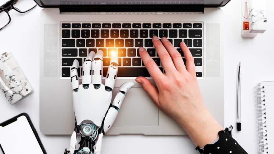 Robotic hand and human hand typing together on a laptop keyboard representing AI-human collaboration in content creation